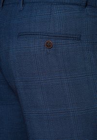 Granatowe spodnie w kratę z teksturowanej tkaniny. Posiadają tylnią kieszeń z brązowym zapięciem na guzik. Subtelny wzór w kratę na całej powierzchni.