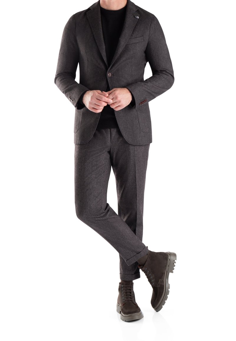 Traje gris a medida de tela texturizada, camisa negra y botas marrones con suelas rugosas. El traje cuenta con solapas de muesca y detalles de botones.