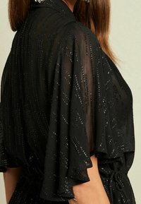Blouse noire de style kimono avec manches fluides, en tissu transparent. Comprend des rayures métalliques et un détail de taille nouée.