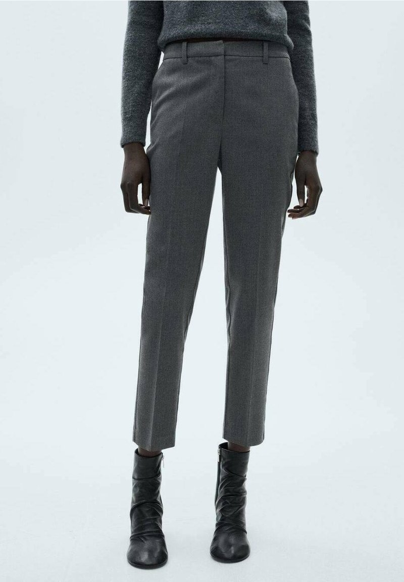 Personne portant un pantalon gris près du corps, un pull gris et des bottines noires, debout devant un fond clair uni.