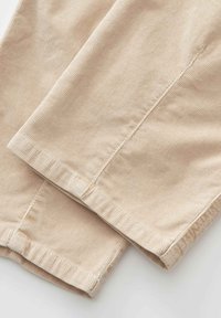 Nahaufnahme einer beige Cordstofftextur, die gefaltete Hosenbeine mit sichtbaren Nähten und Saumdetails zeigt.