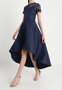 Marineblauwe asymmetrische jurk met kanten korte mouwen, getailleerd lijfje en vloeiende rok. Gemaakt met naakte hoge hakken.