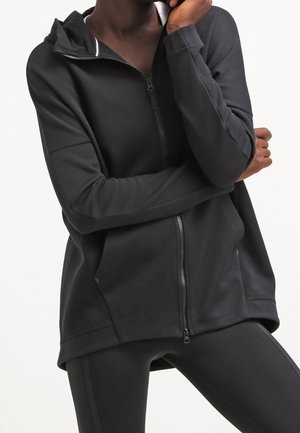 Osoba ubrana w czarną bluzę z kapturem zapinaną na zamek, z asymetrycznym dołem, oraz czarne legginsy, z założonymi rękami, z jedną dłonią dotykającą twarzy.