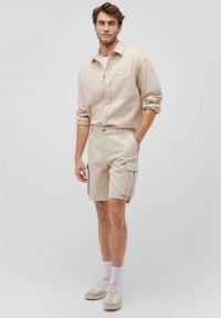 Beige Baumwollhemd mit langen Ärmeln, kombiniert mit passenden Cargo-Shorts, die Taschen und eine lockere Passform haben. Getragen mit weißen Socken und Sneakern.