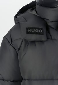 Black padded jacket with a hood featuring a black patch label with the brand name "HUGO" on the shoulder area.