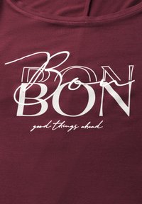 Bordó tričko s velkým bílým potiskem obsahující slovo "BON" a menší text "dobré věci přijdou". Měkká struktura, přiléhavý střih a kulatý výstřih.