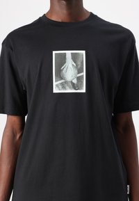 Sort bomulds t-shirt med et centralt grafisk motiv af en hånd, der holder en basketball, med en rektangulær hvid kant omkring billedet.