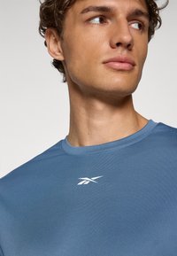 Lyseblå atletisk T-shirt lavet af struktureret stof. Har en rund halsudskæring og et hvidt logo på venstre brystområde.