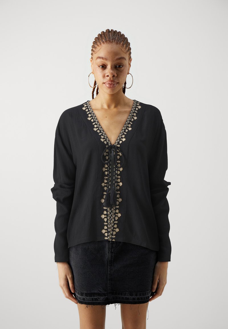 Blusa nera con maniche lunghe e ricami dorati decorativi lungo il colletto. Presenta un scollo a V e una chiusura a fiocco sul davanti.