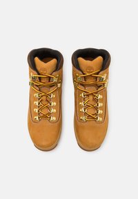 Botas de senderismo de cuero marrón con herrajes dorados, cordones amarillos y rojos, cuello acolchado y un logo en la lengüeta. Diseño de suela plana.
