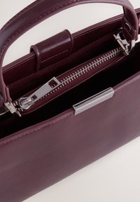 Vinska rdeča usnjena torbica z gladko teksturo, zgornjimi ročaji in srebrnim zadrgo. Ima strukturo zasnove z elegantnim zaključkom.