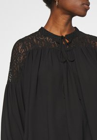 Blouse noire à coupe ample, avec des accents en dentelle noire sur les épaules et une fermeture à nouer au niveau du cou. Tissu doux et fluide.