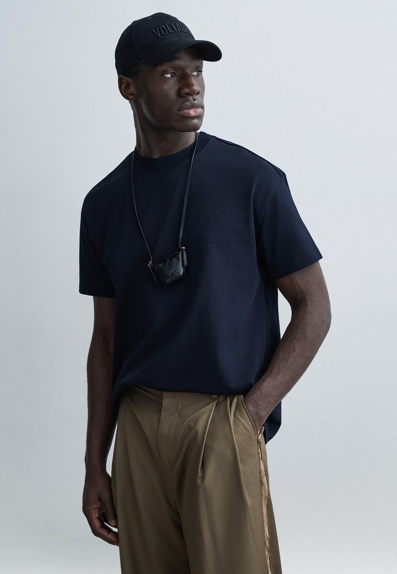 Námořnické tričko s kulatým výstřihem z žebrované látky, doplněné světle hnědými plisovanými kalhotami s bočními pruhy a černou čepicí s nápisem „VOLTAIRE“.