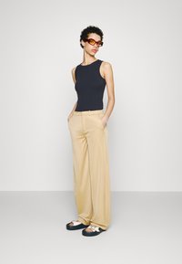 Svart ermeløs topp, beige høytlivede vide bukser og to-fargede sandaler med spenne detaljer. Modellen poserer med hendene i lommene.