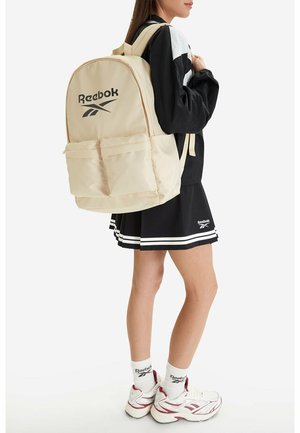 Donna che indossa un completo sportivo bianco e nero con sneakers bianche Reebok, calzini bianchi Reebok e zaino beige Reebok sulla schiena.