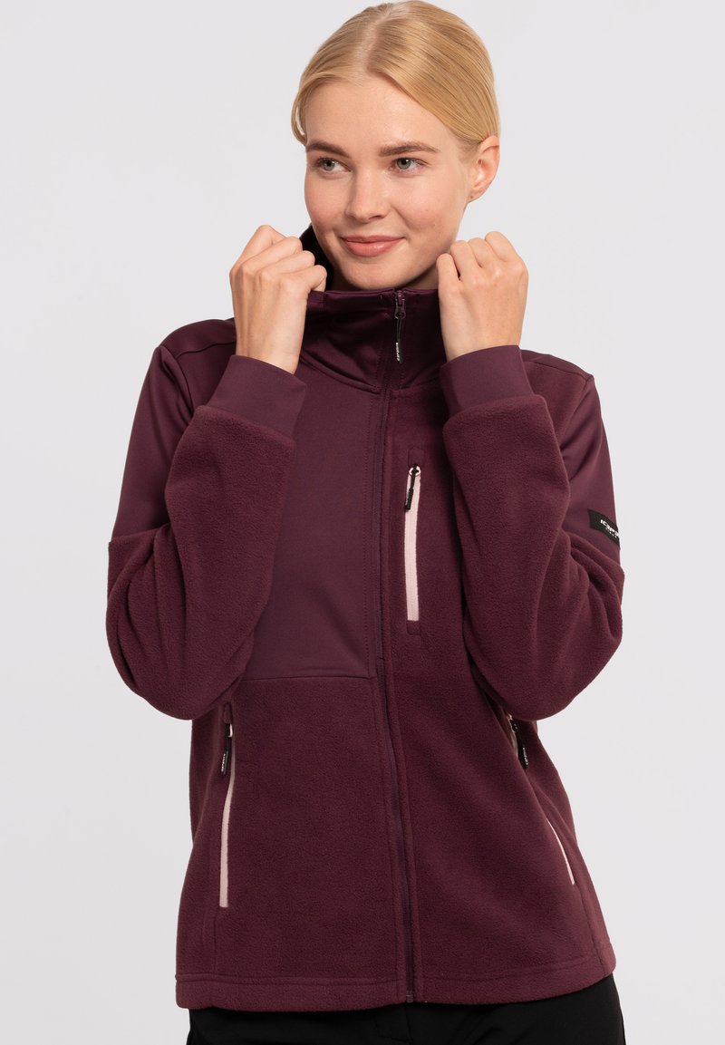 Veste en polaire bordeaux avec un col haut, des poches zippées et un design ajusté. Caractéristiques d'accents clairs contrastants et d'un tissu texturé.