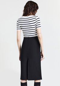 Frau mit nassen, dunklen Haaren, die ein weißes, kurzärmeliges Shirt mit schwarzen Streifen und einen hochtaillierten, schwarzen Midirock trägt, von hinten betrachtet.
