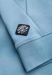 Lyseblå sweatshirtstof med tekstureret finish. Har et sort hexagonalt mærke mærket "OMBRE 1985," broderet detalje og manchetter.
