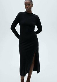 Abito lungo nero a maniche lunghe realizzato in tessuto knit, presenta un collo alto e una fessura laterale, che arriva a metà polpaccio. Texture liscia con una vestibilità aderente.