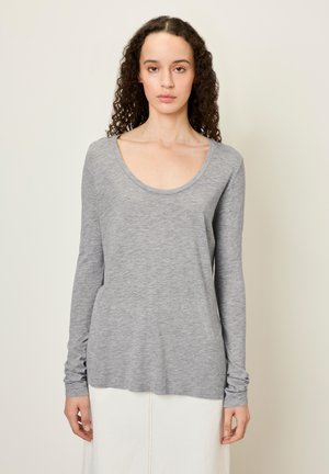 Woman with curly hair wearing a light gray long-sleeve top and white skirt, standing against a plain beige background.