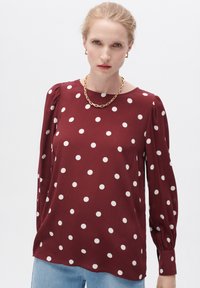 Bordeaux blouse met witte stippen, lange mouwen met geplooide schouders, ronde halslijn en een gladde stoftextuur.