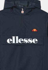 Granatowa kurtka przeciwwiatrowa z designem na pół zamka, z meshowym wykończeniem kaptura, dużą kieszenią z przodu oraz białym logo "ellesse". Zawiera małą naszywkę z logo.