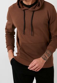 Sudadera marrón hecha de una tela suave, con textura acanalada en las mangas y la capucha, bolsillo frontal y cordones negros.