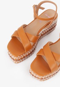 Tan leather platform sandals with crossed straps, square toe, and a woven texture on the wedge sole featuring white stitching details.