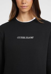 Felpa nera realizzata in misto cotone; presenta un collo a rondine e un logo bianco "GUESS JEANS" ricamato sul davanti. Texture liscia.