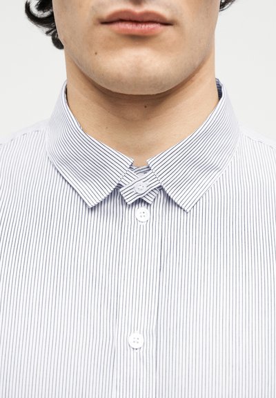 Κοντινό πλάνο του κάτω μέρους του προσώπου και του λαιμού ενός ατόμου που φοράει άσπρο πουκάμισο με λεπτές κατακόρυφες μαύρες ρίγες, κουμπωμένο στο πάνω μέρος.