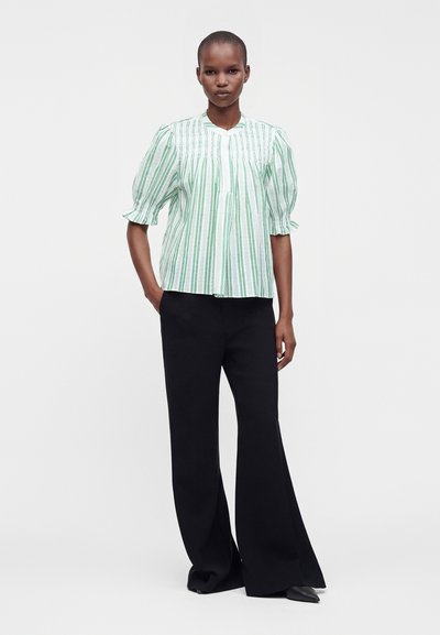 Femme portant un chemisier à manches bouffantes rayé vert et blanc et un pantalon noir à jambes larges, debout avec une main dans la poche sur fond blanc.