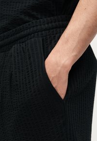 Χέρι που ακουμπάει μέσα στην υφασμάτινη τσέπη με υφή μαύρου χρώματος ενός ενδύματος, εμφανίζοντας τον καρπό και μέρος του αντιβραχίου.