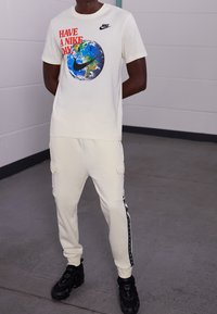 Cream short-sleeve T-shirt with a globe and red text front design; paired with cream cargo pants featuring side pockets and black accents. Black sneakers.