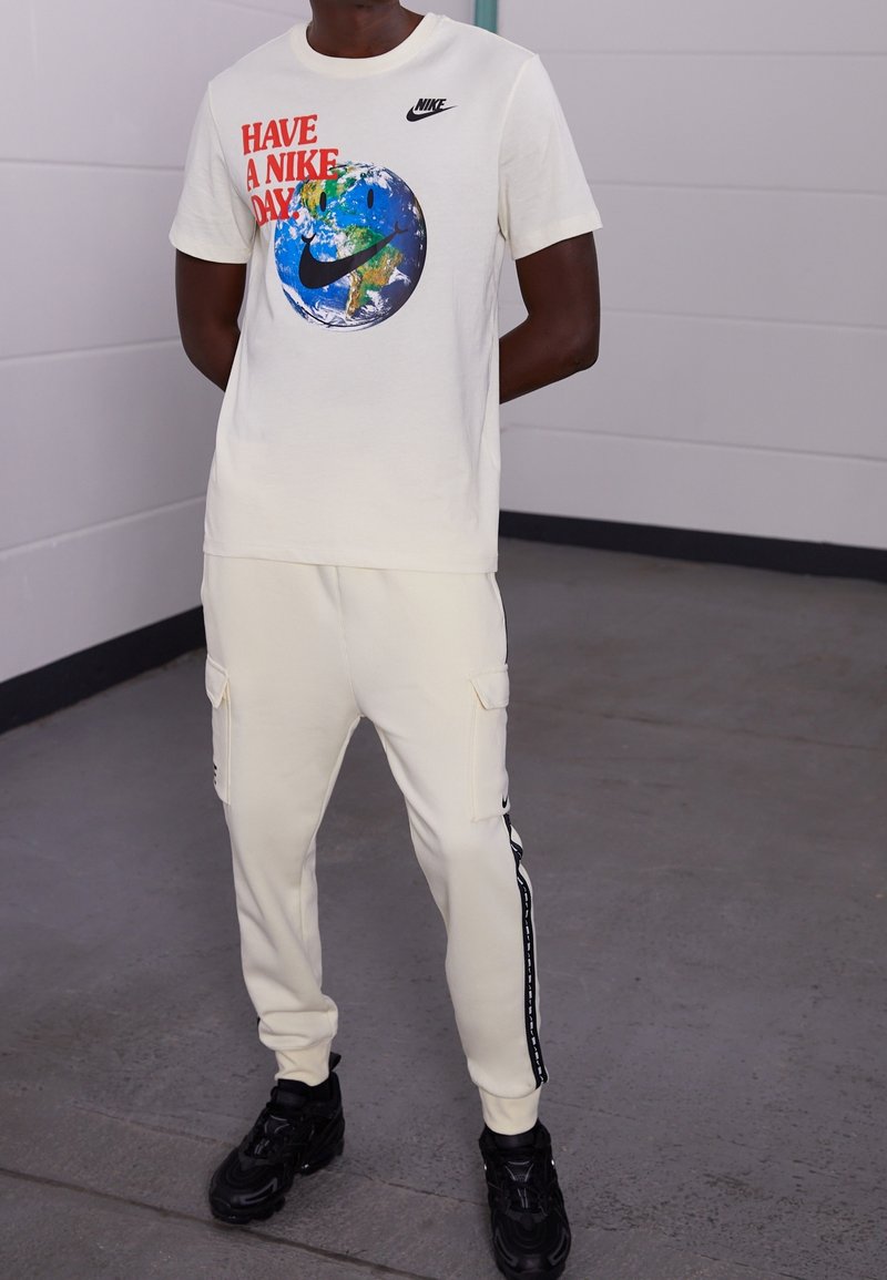 Cream short-sleeve T-shirt with a globe and red text front design; paired with cream cargo pants featuring side pockets and black accents. Black sneakers.