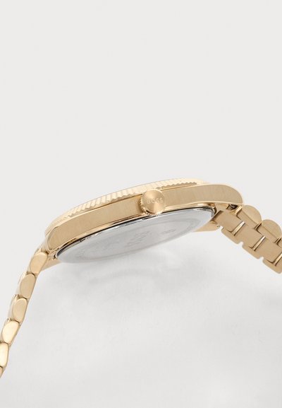 Vue de côté d'une montre-bracelet en or avec un bracelet en métal à maillons et une couronne étiquetée "BOSS" sur un fond clair uni.