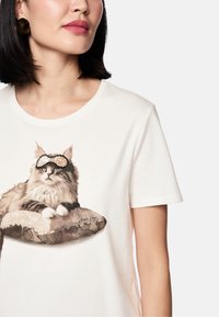 Weißes T-Shirt mit kurzen Ärmeln, das eine Grafik einer Katze zeigt, die eine Sonnenbrille trägt und auf einem dekorativen Kissen mit einem Muster liegt.
