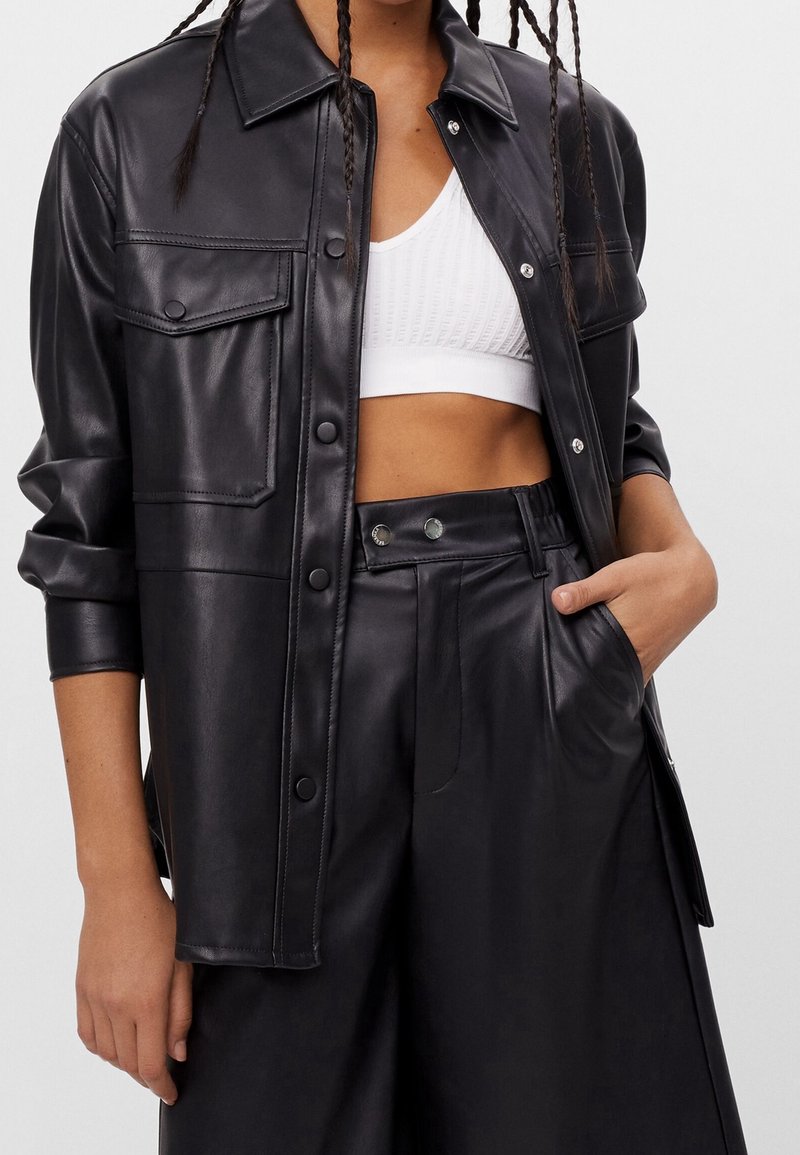 Kvinna i en svart fusk läder skjortjacka, matchande hög midja byxor och en vit kortärmad texturerad tank top, med en hand i fickan.