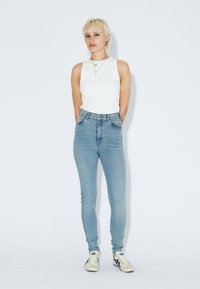 Persona con cabello rubio corto que lleva una camiseta sin mangas blanca, jeans ajustados de tiro alto en un tono azul claro y zapatillas blancas, de pie contra un fondo blanco.