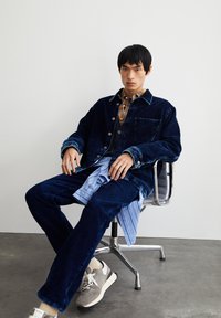 Een jonge man zit op een draaistoel, gekleed in een marineblauwe corduroy jasje en broek, gelaagde shirts en grijze sneakers op een betonnen vloer.