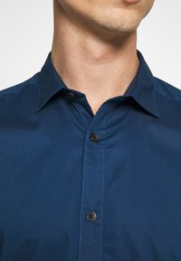 Camisa azul oscuro abotonada con cuello puntiagudo, dos botones frontales y textura suave con costuras contrastantes a lo largo de las uniones.