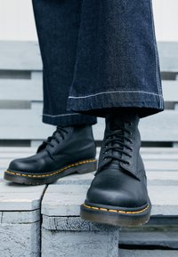 Botas de cuero negro con costuras amarillas, puntera redonda y diseño de cordones, combinadas con pantalones de mezclilla oscura y anchos que descansan sobre una superficie de madera gris.
