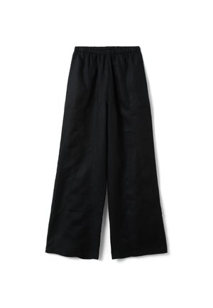 Donna che indossa pantaloni neri a vita alta e gamba larga e scarpe nere a punta, in piedi davanti a un muro grigio con texture.