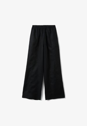 Donna che indossa pantaloni neri a vita alta e gamba larga e scarpe nere a punta, in piedi davanti a un muro grigio con texture.