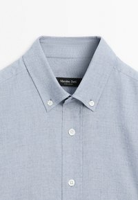 Chemise bleu clair à boutons blancs avec un col pointu, présentant une étiquette de marque visible à l'intérieur du col.