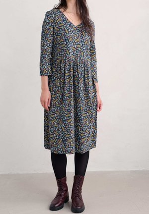 Woman wearing a knee-length floral dress with three-quarter sleeves, black tights, and dark red lace-up boots on a plain background.