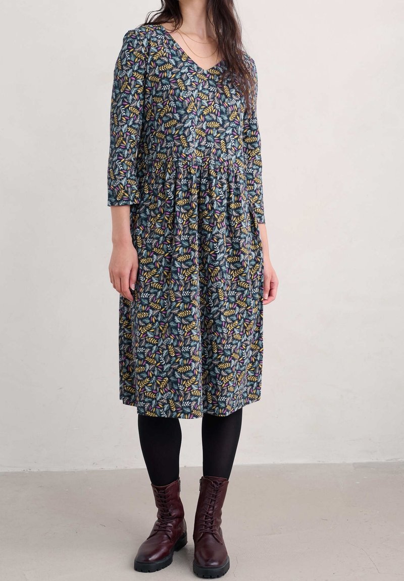 Femme portant une robe à fleurs mi-longue avec des manches trois-quarts, des collants noirs et des bottines à lacets rouge foncé sur un fond uni.