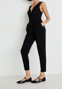 Kobieta w czarnym kombinezonie bez rękawów z dekoltem w serek, z rękami w kieszeniach i czarnych butach Mary Jane, stojąca przy białej ścianie.