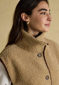 Beige Fleeceveste mit hohem Kragen, braunen Akzenten und Knopfverschlüssen. Der Stoff wirkt strukturiert und weich.