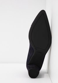 Chaussure à talon noire à bout pointu reposant sur le côté, montrant une semelle lisse avec des marques de taille et de marque.