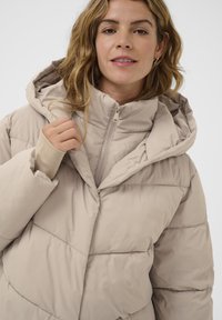 Beige puffer-takki, jossa on suuri huppu, vetoketjulla suljettava etuosa ja joustavat hihansuut. Sen pinnassa on päällyskerros ja ommeltuja yksityiskohtia.